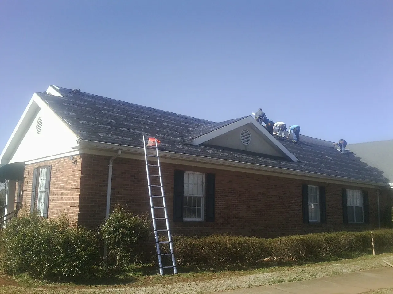 Crew tearing shingles off a brick ranch roof with a ladder set — Emergency Roof Repair work by our crew in Washington Grove