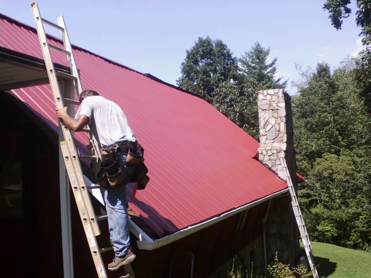 Roofer on a ladder beside a red metal roof and stone chimney — Soffit & Fascia Repair by Roofing Brothers in Washington Grove